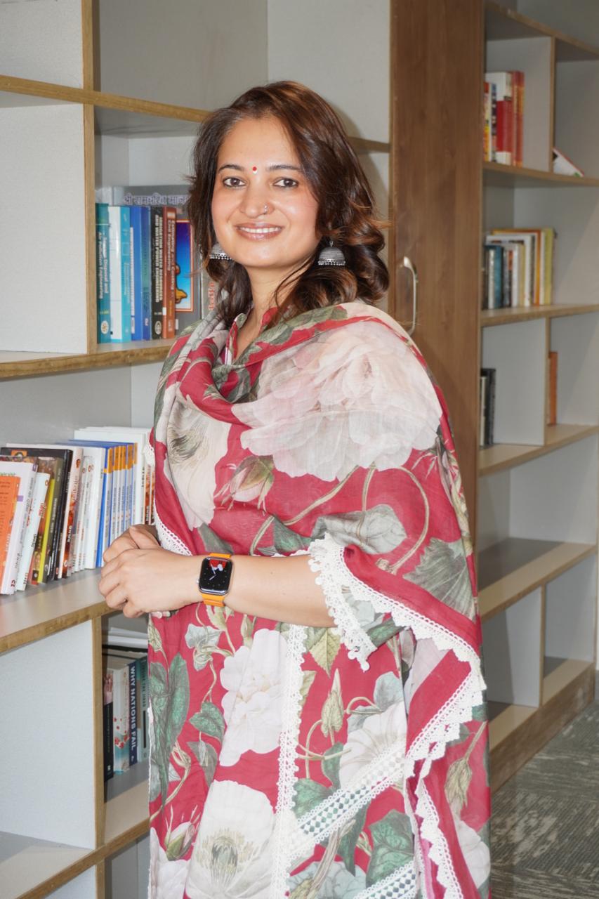 Dr. Sulbha Arya smiling, standing by a bookshelf in a floral saree